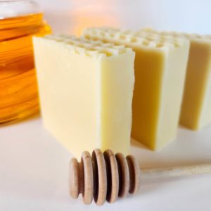 A picture of three bars of soap with a glass jar of honey behind them and a honey dipper in front.