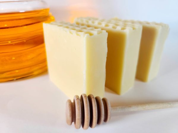 A picture of three bars of soap with a glass jar of honey behind them and a honey dipper in front.