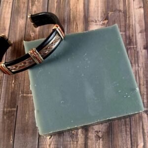 A rugged gunmetal gray bar of soap resting on a wooden background with a masculine bracelet draped over a corner