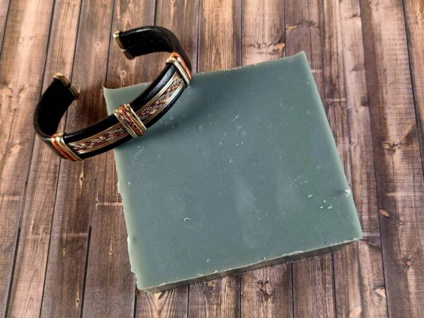 A rugged gunmetal gray bar of soap resting on a wooden background with a masculine bracelet draped over a corner