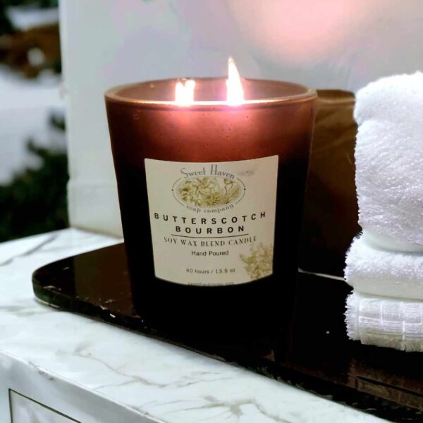 A lit double-wick candle in a matte black jar sitting on a merble countertop next to 4 folded white towels.