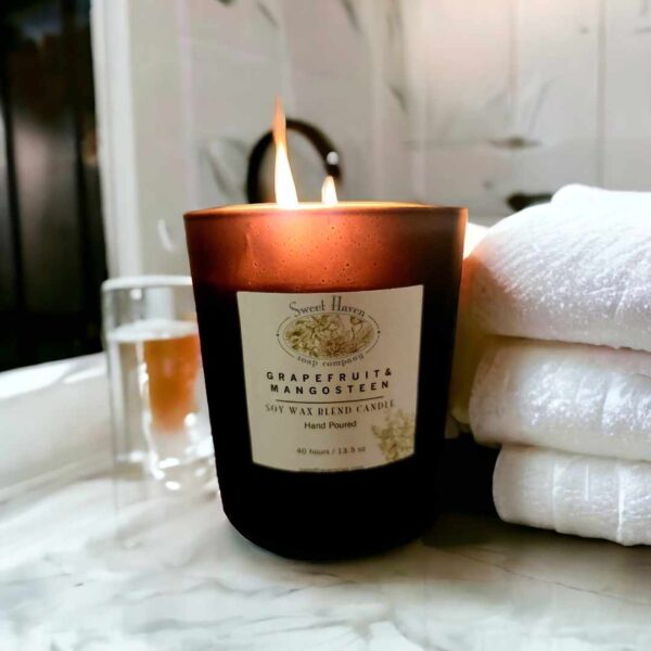 A lit double-wicked candle in a black jar on a white marble bathroom countertop with three folded white towels next to it.