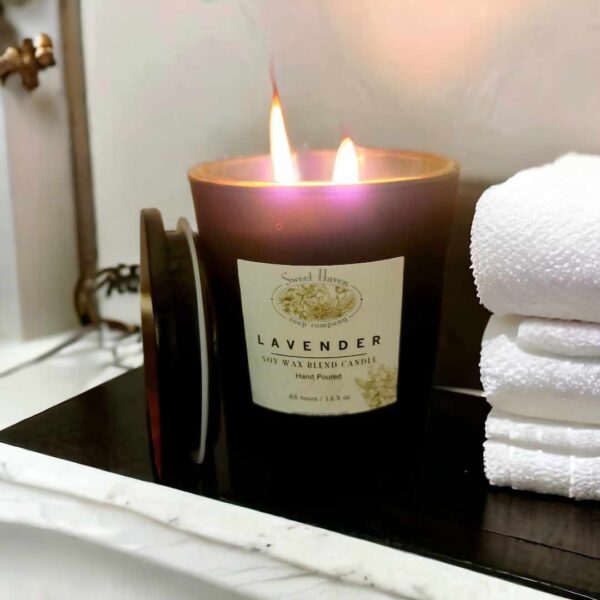 A lit double-wick candle in a black container on a white marble countertop next to 3 thick, folded luxurious towels.