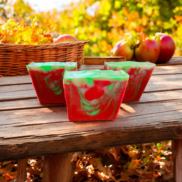 Three red, white, and green swirled soaps setting on a wooden table with apples in the background during a sunny Fall Day.