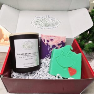 A red Christmas gift box with a candle in a matte black glass container, a purple, pink, and white swirled soap, and a green soap with the Grinch stamped on it.